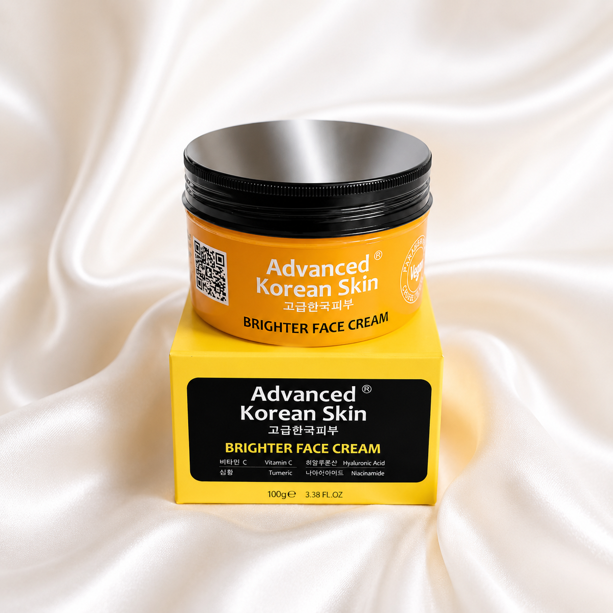 A jar of Advanced Korean Skin Brighter Face Cream sits on top of its yellow retail box against a draped white satin background. The jar has a black lid and a yellow label, while the box features a black front panel with white and yellow text.