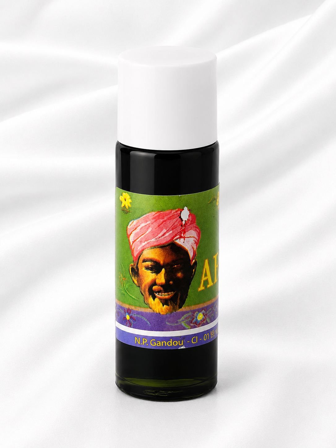 A small cylindrical bottle with a white cap and a colorful illustrated label, photographed against a white draped fabric background. The label shows a portrait with a pink turban on a green and purple background.