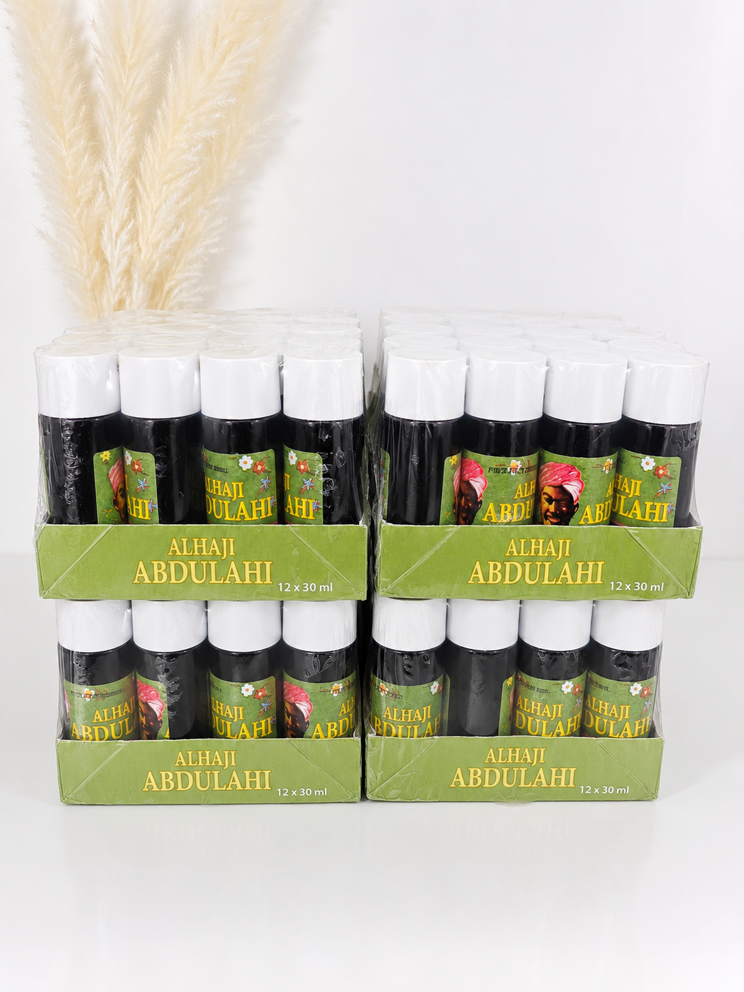 Two cardboard display trays holding multiple small bottles labeled 'ALHAI ABDULAHI' on a white background, with dried pampas grass decor behind them.