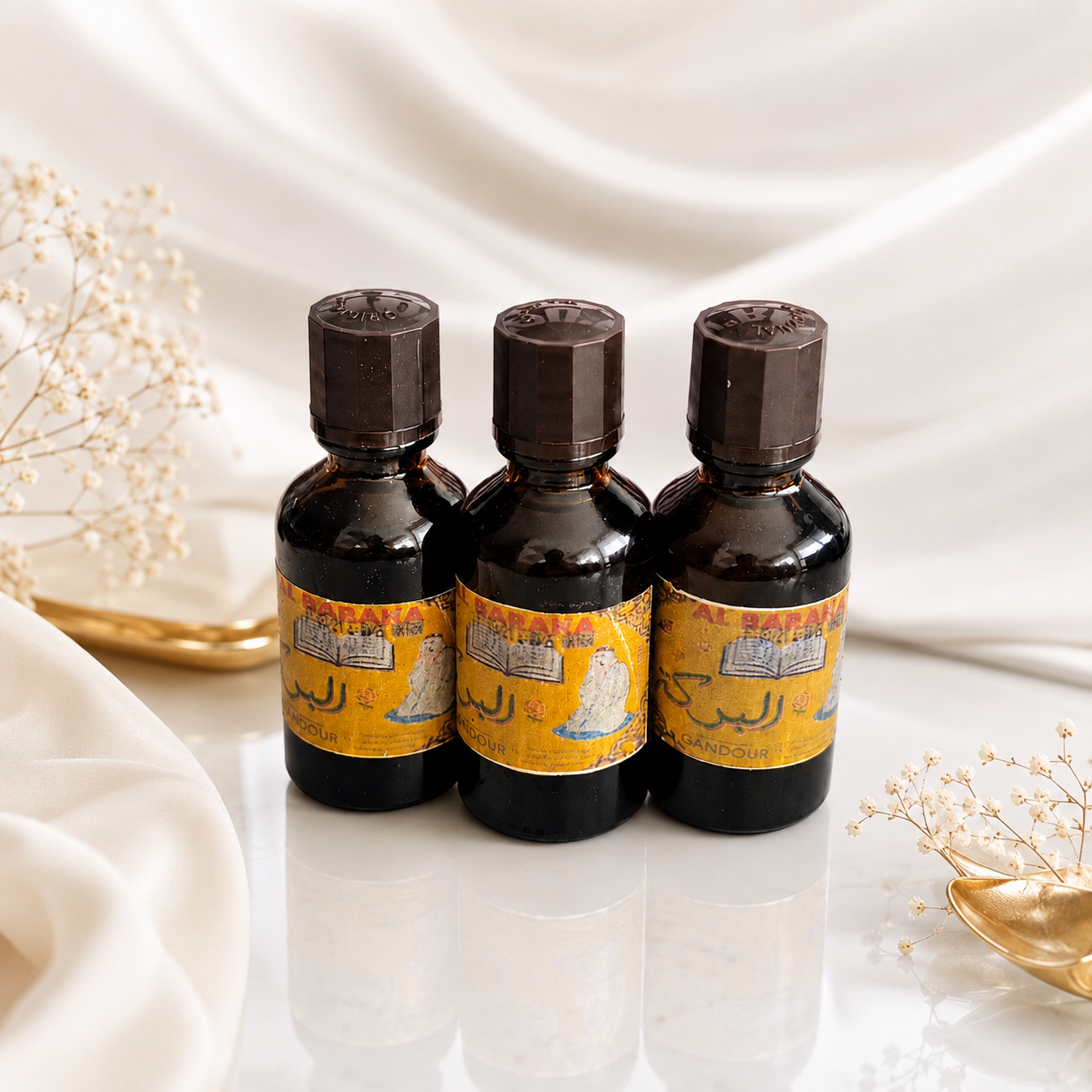 Three small dark amber bottles with yellow labels and dark caps, arranged together on a glossy white surface with draped white fabric and small dried floral props in the background.