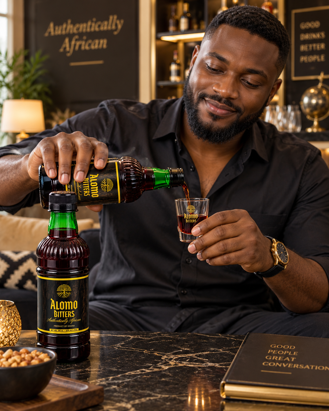 A man in a black shirt pours Alomo Bitters from a green bottle into a small glass, with another bottle and a bowl of peanuts on a marble table in a warm, upscale bar setting.