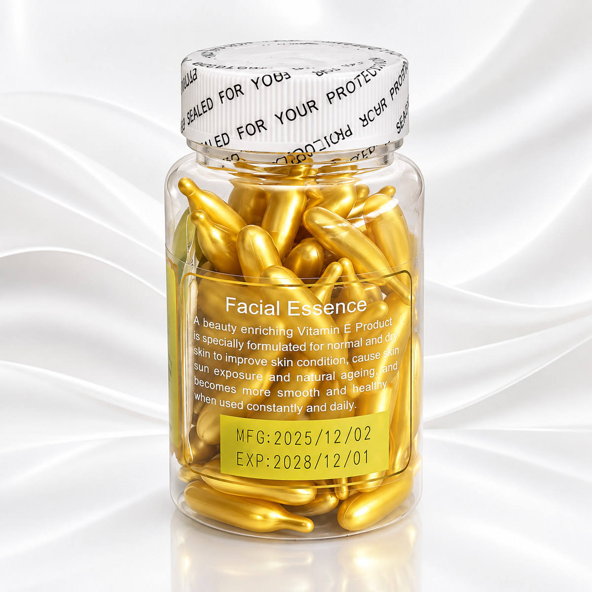 A clear glass bottle filled with shiny gold capsules, sealed with a white branded cap, on a white satin-textured background. The front label reads 'Facial Essence' with additional product text and dates.