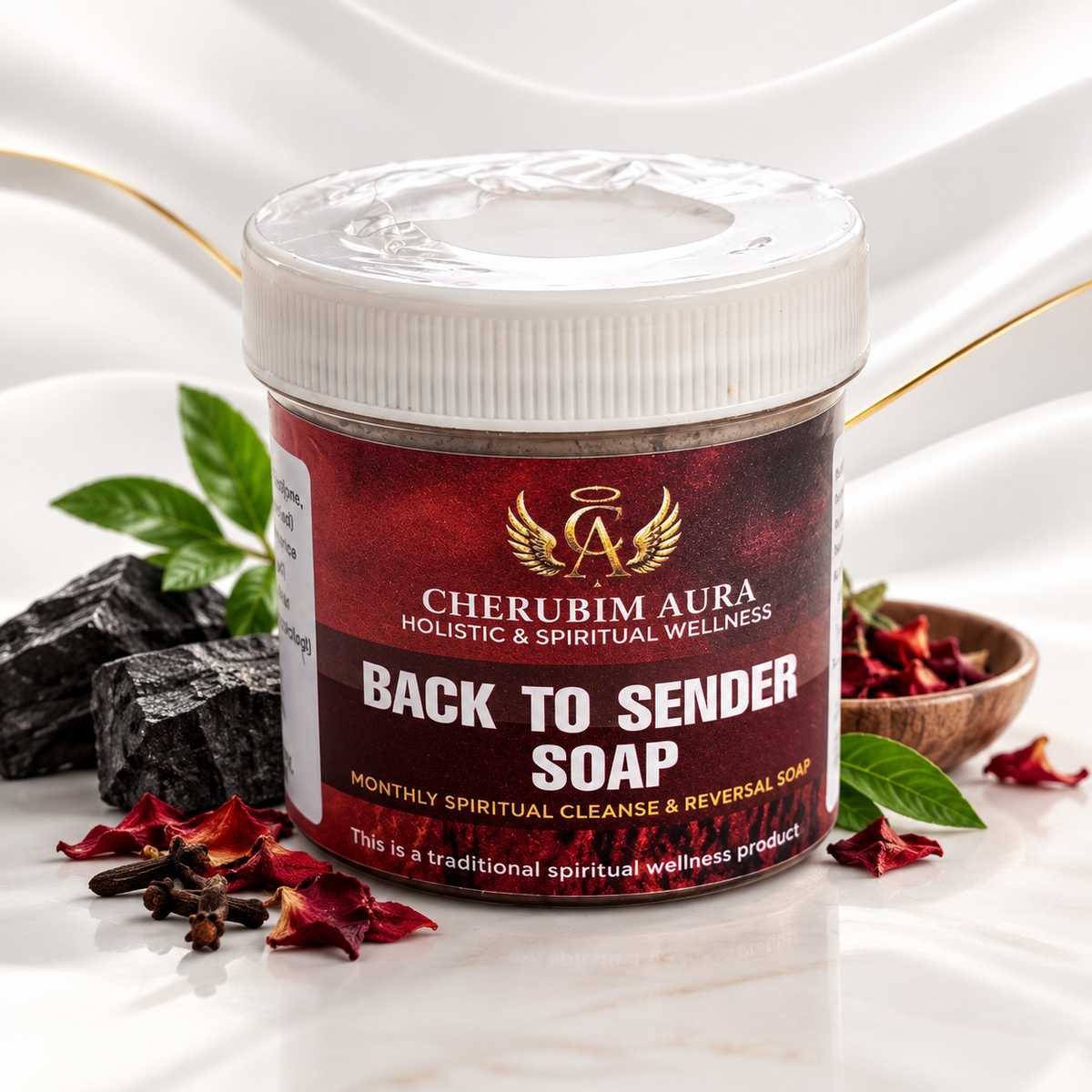 A jar of Cherubim Aura Back to Sender Soap on a white marble surface with red rose petals, black stones, and green leaves. The product is a clear plastic jar with a white screw cap and a dark red label with gold and white text.