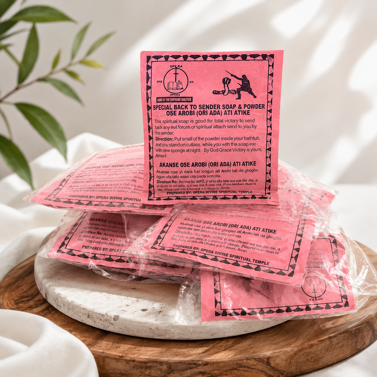 Pink ritual soap packets wrapped in clear plastic, with one packet standing upright to show the printed pink label. The packets are stacked on a round wooden tray with white fabric and green leaves in the background.