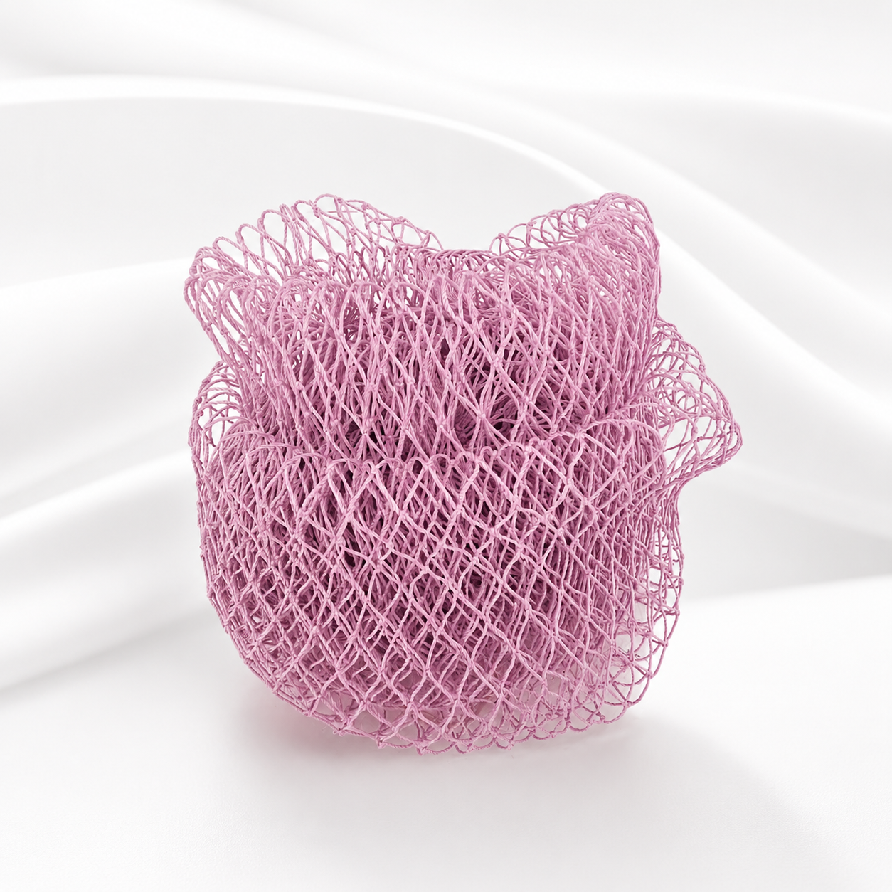 A pink mesh or net-like decorative object with a rounded, organic shape and irregular edges, photographed on a clean white background with soft draped fabric behind it.