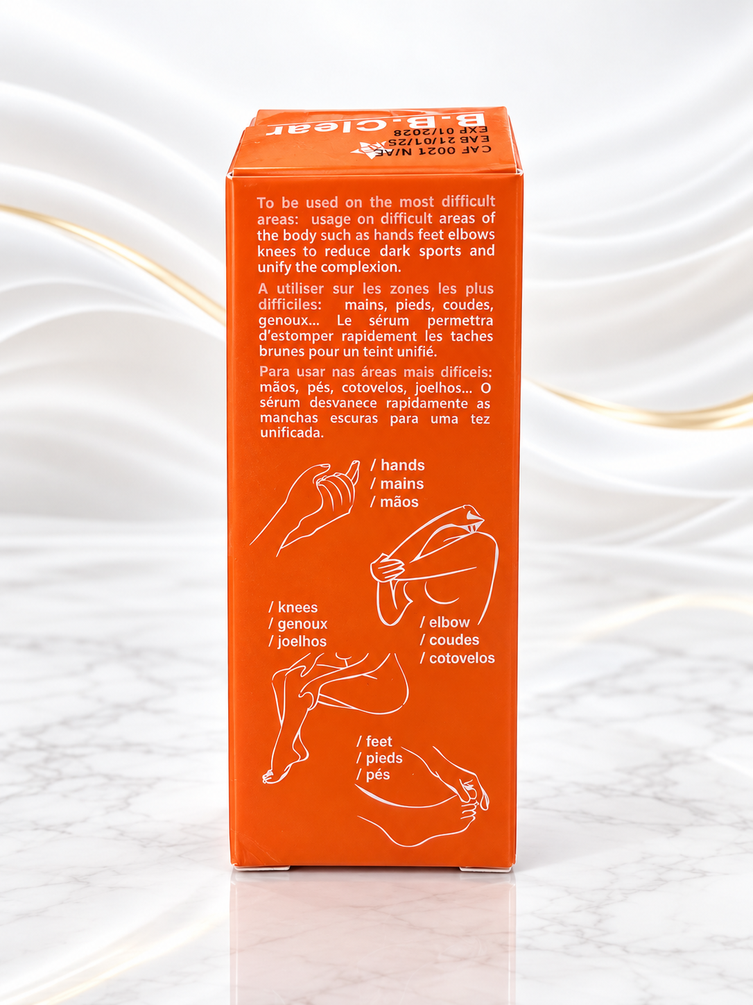 An orange rectangular product box standing upright on a white marble surface with a white draped fabric backdrop. The box shows usage instructions and line drawings in French and Portuguese indicating how to apply the product to hands, knees, elbow, and feet.
