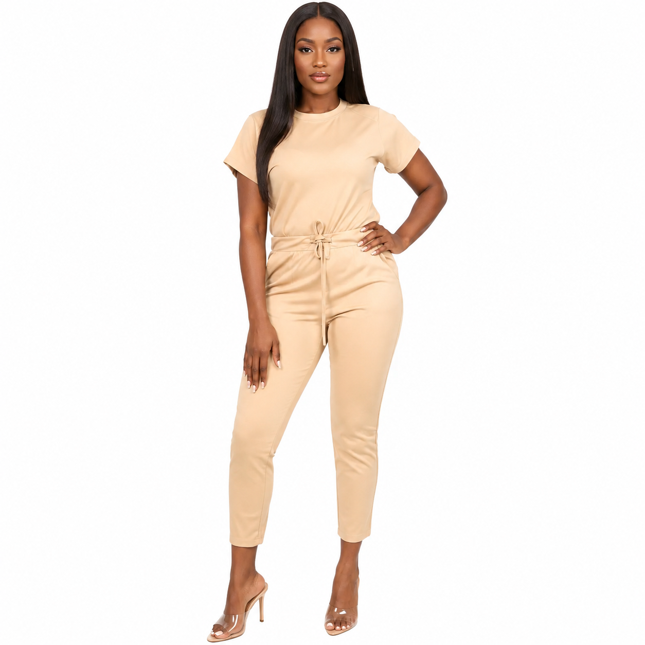 A woman modeling a light beige short-sleeve jumpsuit with a drawstring waist, cropped ankle-length legs, and clear high-heeled sandals against a plain white background.