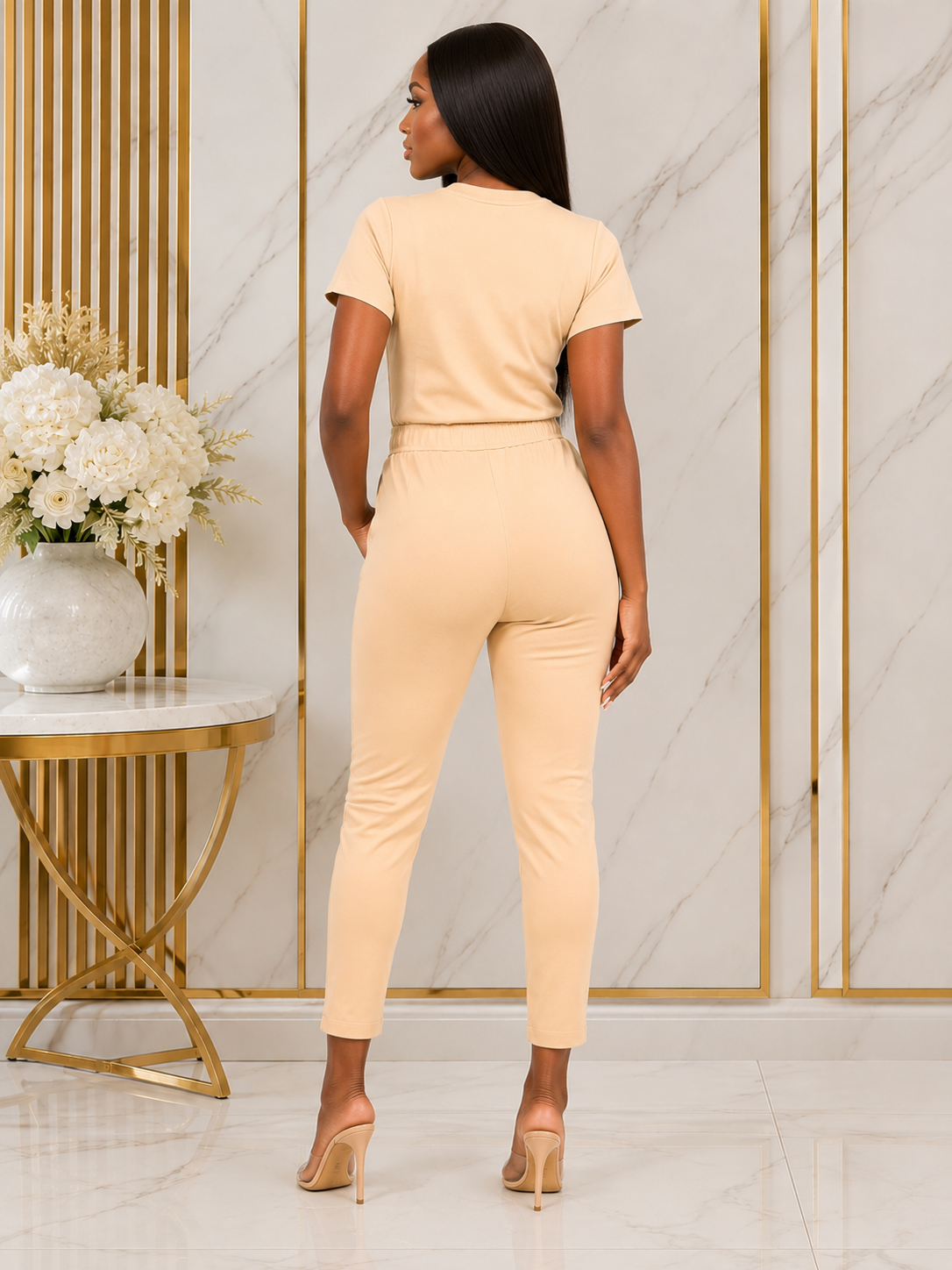 A woman shown from the back wearing a light peach short-sleeve crop top and matching high-waisted cropped pants, standing in a modern interior with marble walls and gold trim.