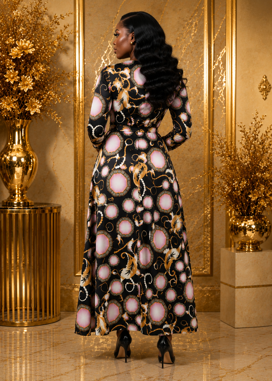 A woman stands with her back to the camera wearing a long black maxi dress with a pink and gold abstract pattern, set against a luxurious gold-toned interior background.