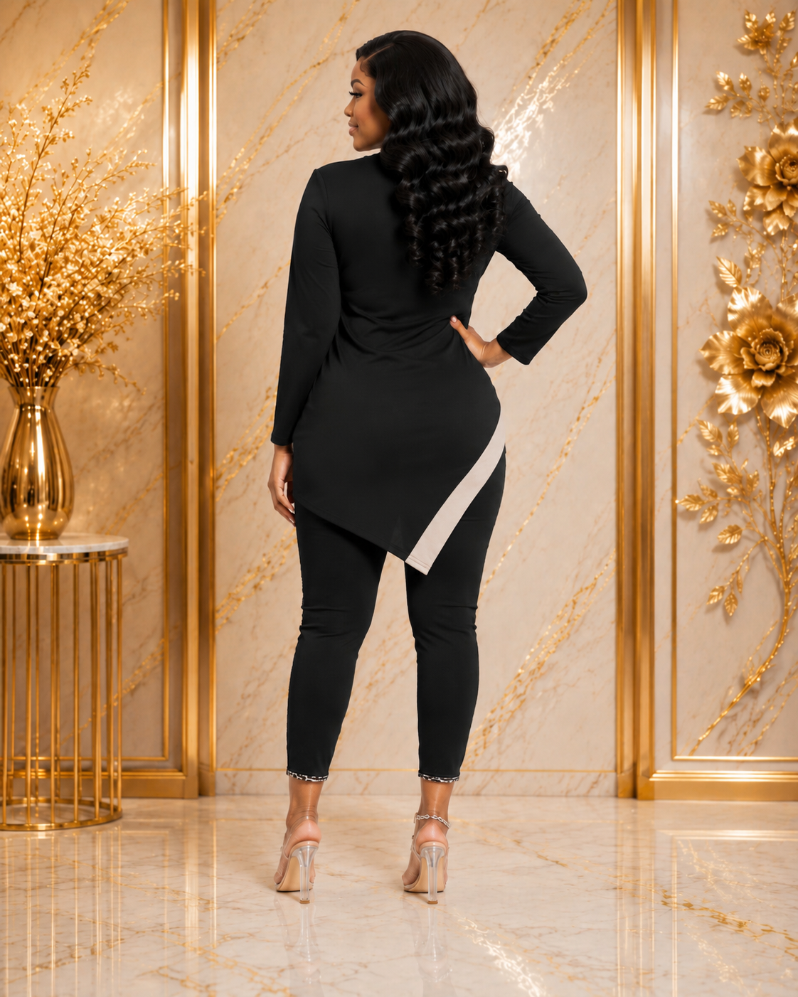 A woman is shown from the back wearing a black long-sleeve tunic top with an asymmetrical hem and a contrasting white side panel, paired with black leggings and nude high heels, set against a luxurious gold and beige marble wall background.