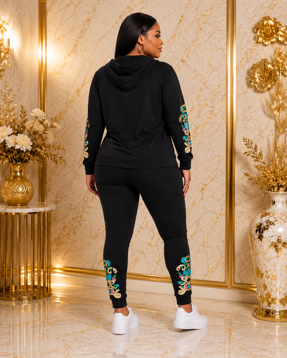 A woman wearing a black two-piece tracksuit with colorful floral embroidery on the sleeves and legs, shown from the back in a luxurious room with gold walls and marble floors.