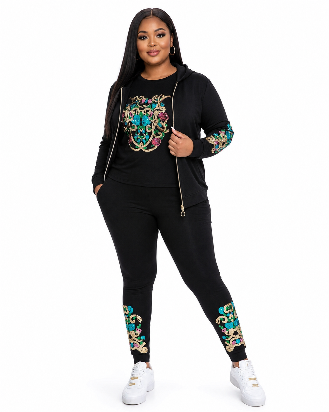 A woman modeling a black two-piece tracksuit with colorful baroque-style embroidery on the chest, sleeves, and lower legs, paired with white sneakers against a plain white background.