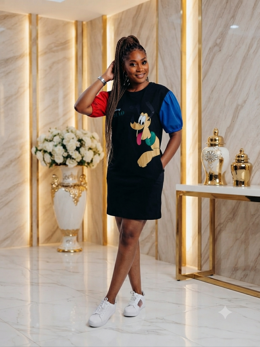 A woman modeling a black short-sleeve dress with a colorful cartoon character graphic on the front, red and blue color-blocked sleeves, and white sneakers, standing in a luxurious marble hallway with gold accents.