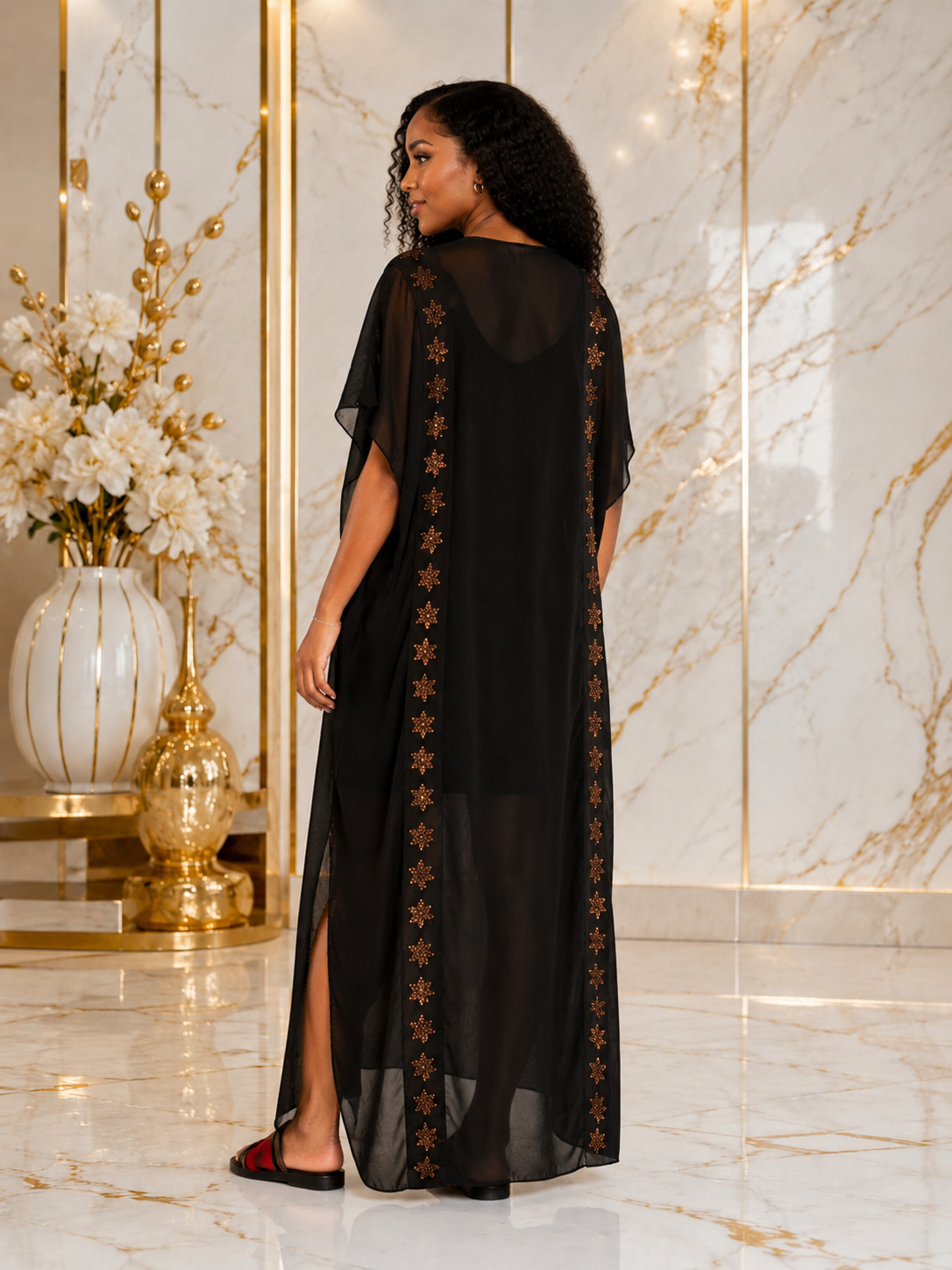 A woman wearing a long black sheer kaftan dress with gold embroidered star patterns along the side panels, standing in a luxurious marble hallway with gold accents.