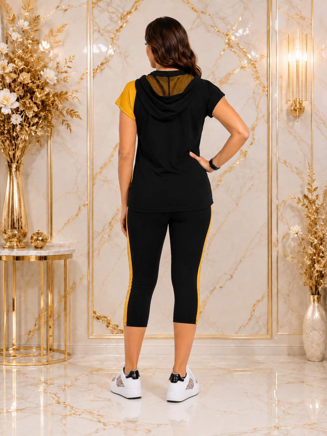A woman viewed from the back wearing a black athletic set with yellow accents, consisting of a short-sleeve hooded top and matching capri leggings, in a luxury interior with gold marble walls and decorative gold vases.