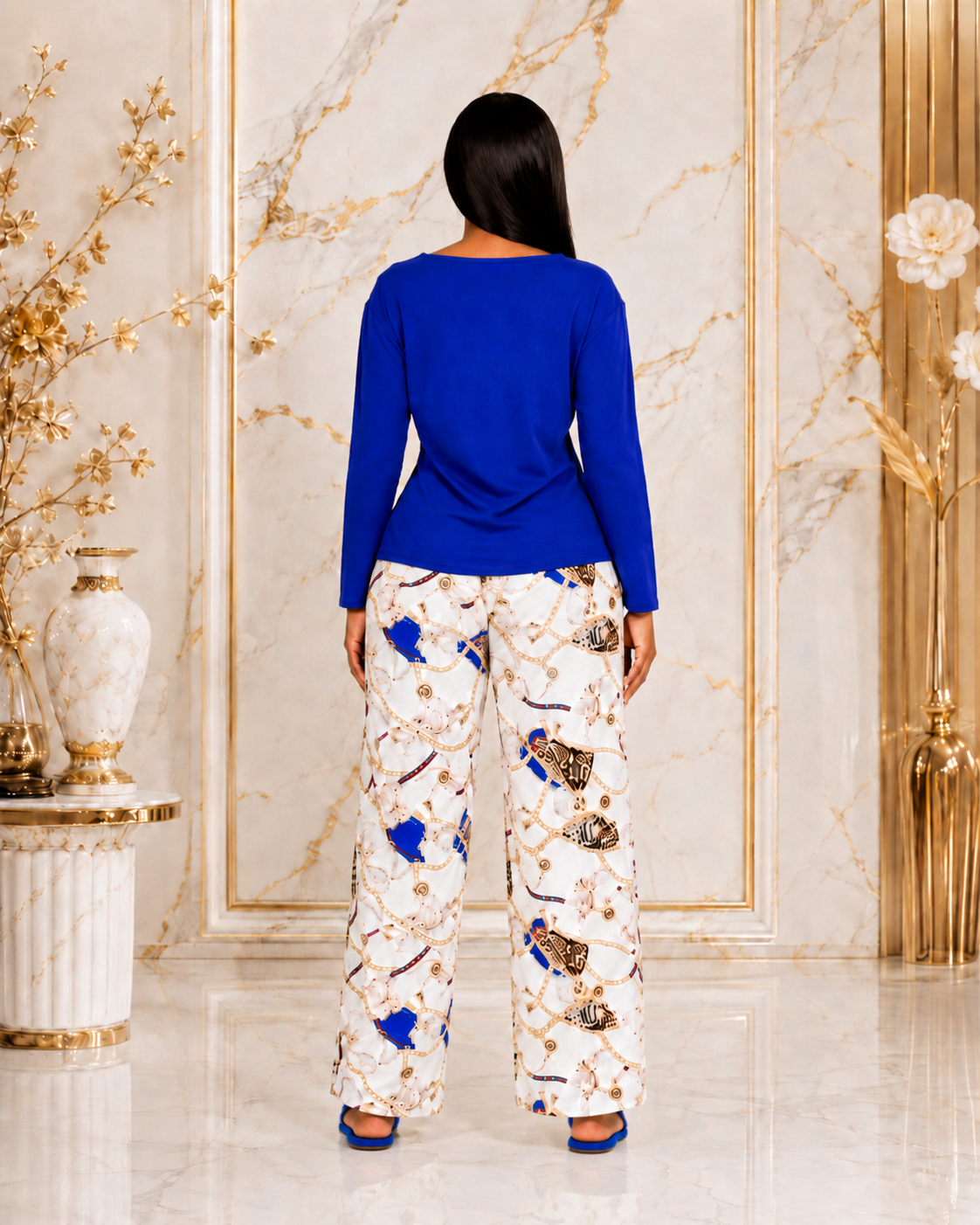 A woman viewed from behind wearing a royal blue long-sleeve top and white wide-leg pants with a blue, gold, and brown abstract print, standing in a luxurious marble-walled room.