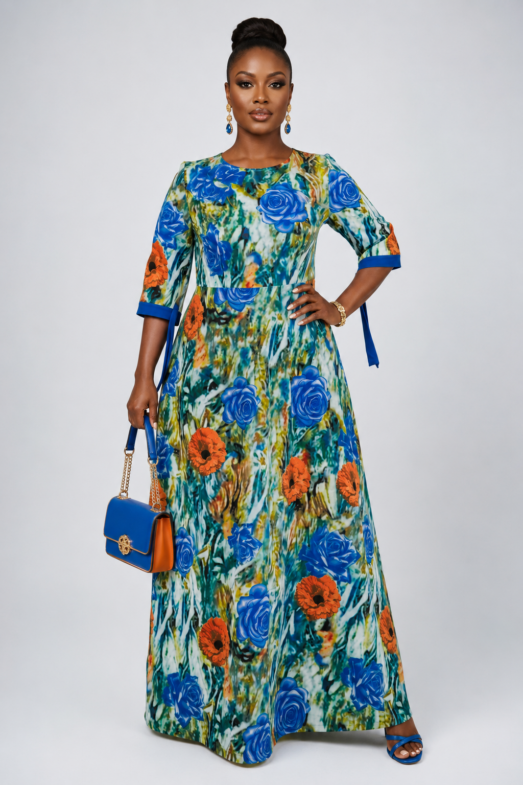 A woman modeling a long-sleeved maxi dress with a vibrant blue, orange, and green floral print, holding a blue handbag against a light gray studio background.