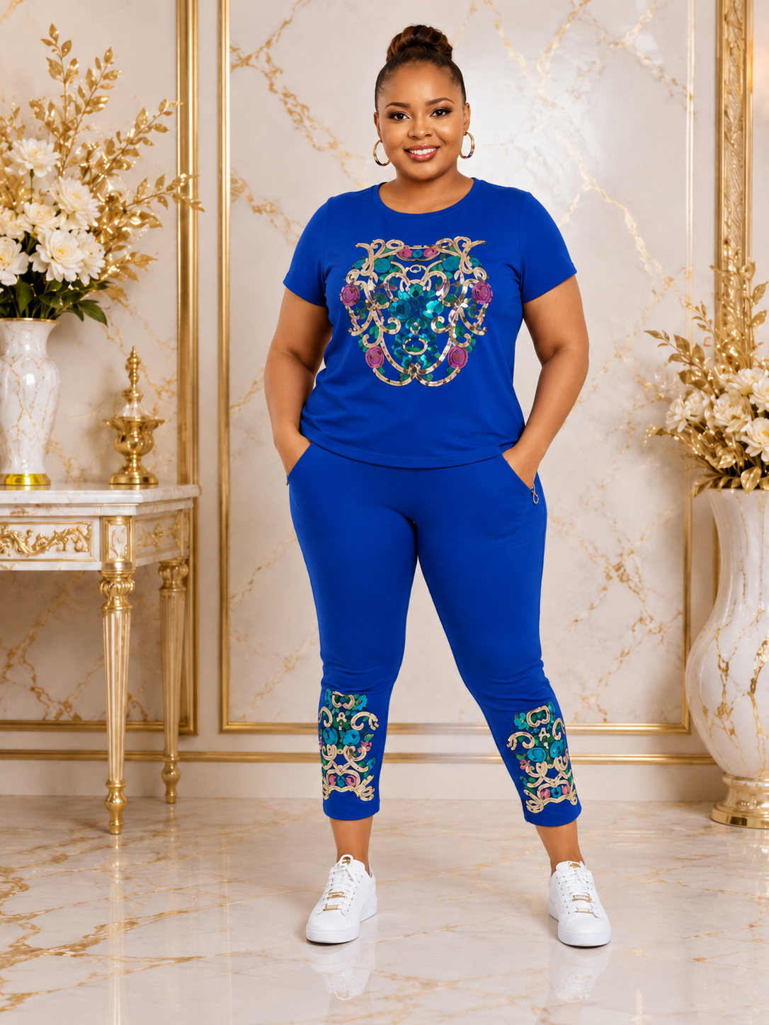 A woman modeling a matching royal blue lounge set consisting of a short-sleeve t-shirt and jogger pants, both featuring ornate multicolor embroidered patterns. She is standing in a luxurious room with gold-trimmed marble walls and white sneakers.