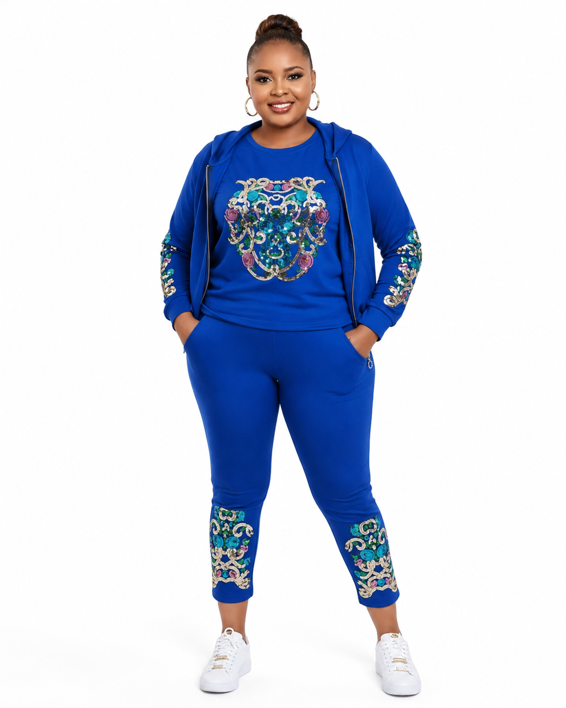 A woman modeling a matching royal blue two-piece tracksuit with colorful embroidered details on the chest and sleeves, paired with white sneakers against a plain white background.