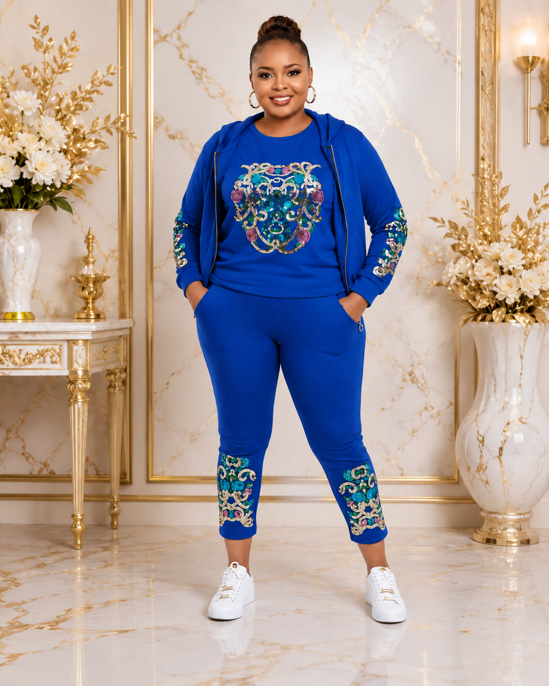A woman modeling a royal blue two-piece lounge set with colorful embroidered details, standing in a luxurious room with gold-trimmed marble walls and white floral arrangements.