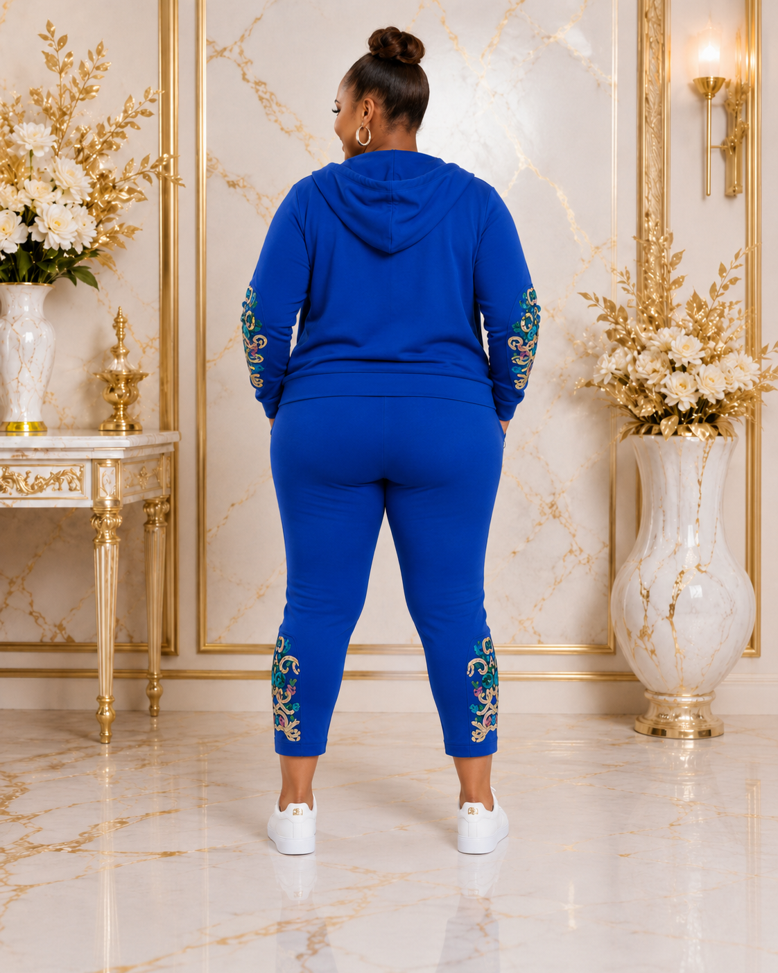 A woman stands with her back to the camera wearing a royal blue matching lounge set with gold and green floral embroidery on the sleeves and lower legs, set in an elegant room with gold-framed walls and marble floors.