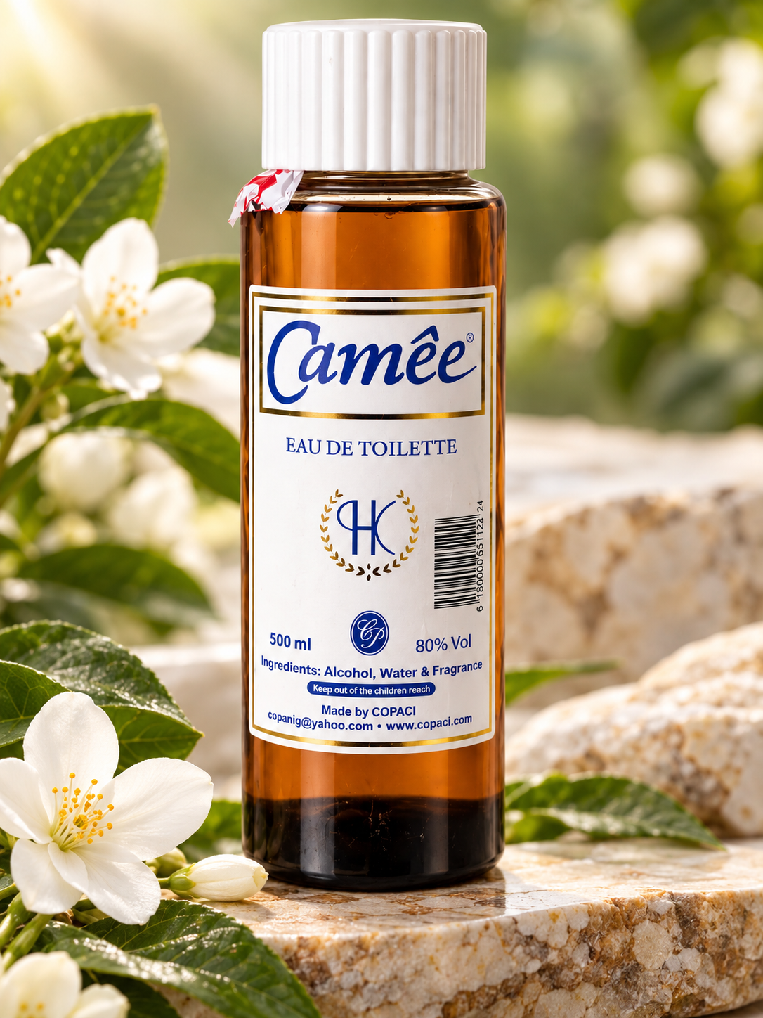 A 500 ml amber glass bottle of Camée Eau de Toilette with a white cap, set on a stone surface surrounded by white flowers and green leaves.