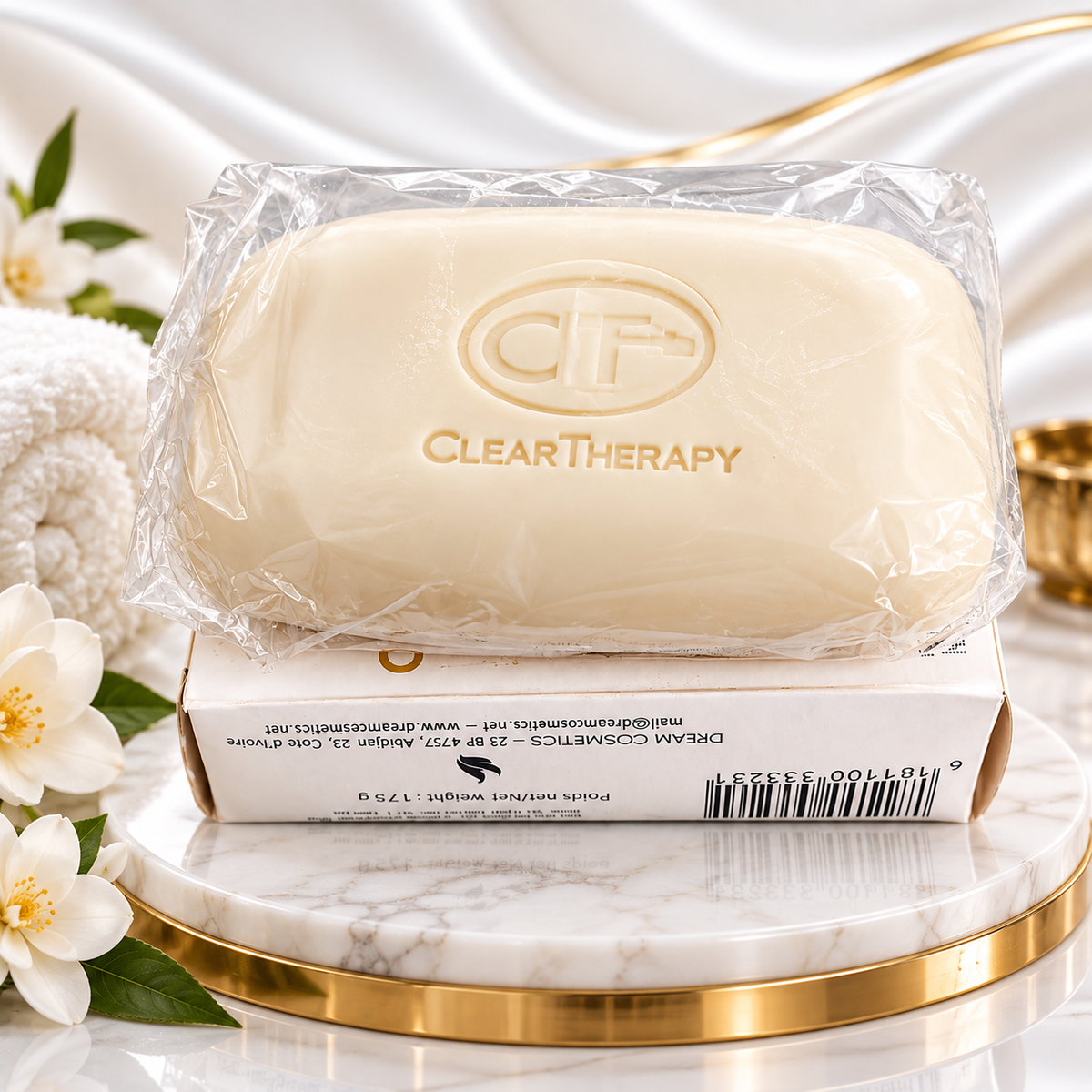 A ClearTherapy cream-colored soap bar wrapped in clear plastic, displayed on top of its white and gold box on a marble pedestal with white flowers and a satin backdrop.