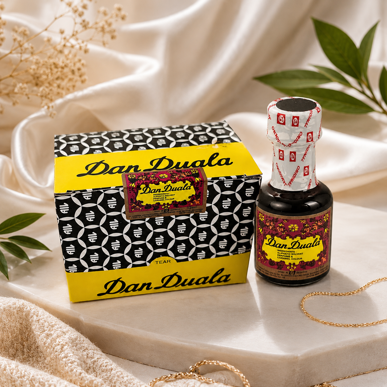 Dan Duala perfume bottle and its decorative packaging box, styled on a white marble tray with draped cream fabric, dried flowers, and green leaves.