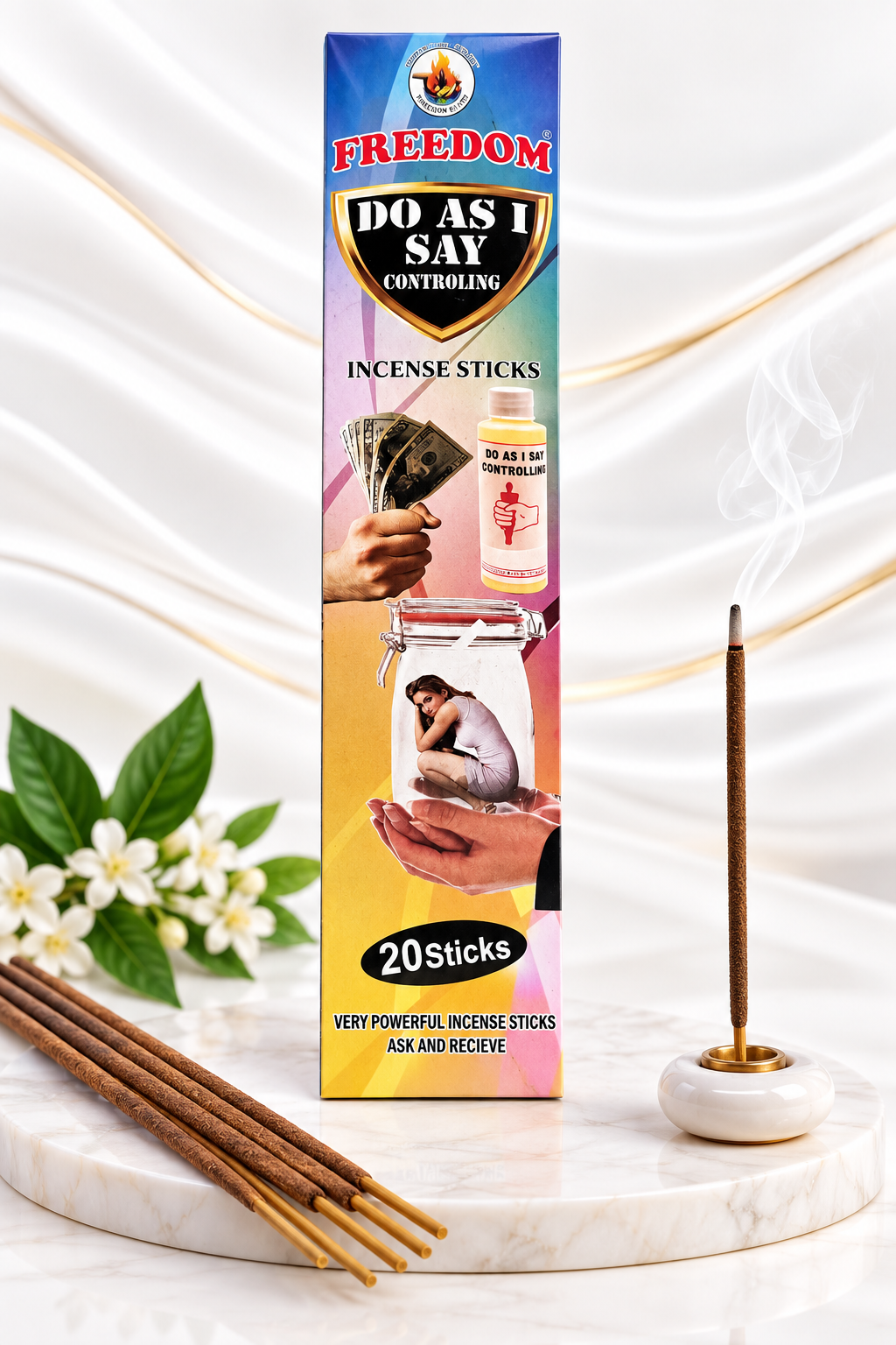 A boxed incense stick product labeled 'FREEDOM DO AS I SAY CONTROLLING' stands upright on a marble surface, with several incense sticks placed in front and a single burning incense stick in a white holder to the right. The background is a soft white draped fabric with subtle gold veining and a few white flowers and green leaves at the lower left.