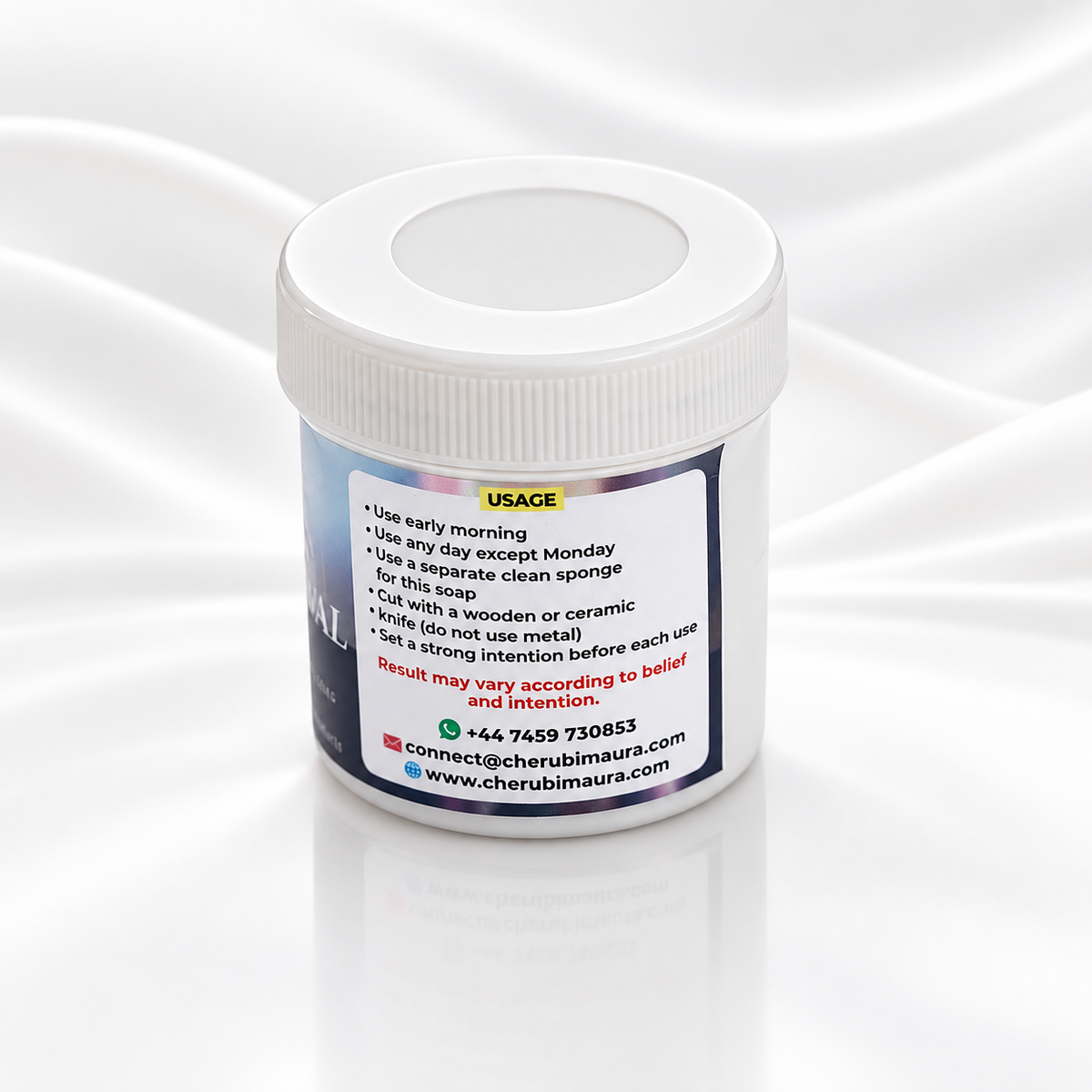 A white plastic jar with a ribbed screw-on lid, shown on a glossy white reflective surface with a soft draped satin fabric background. The front of the jar features a white label with instructions under the heading 'USAGE'.