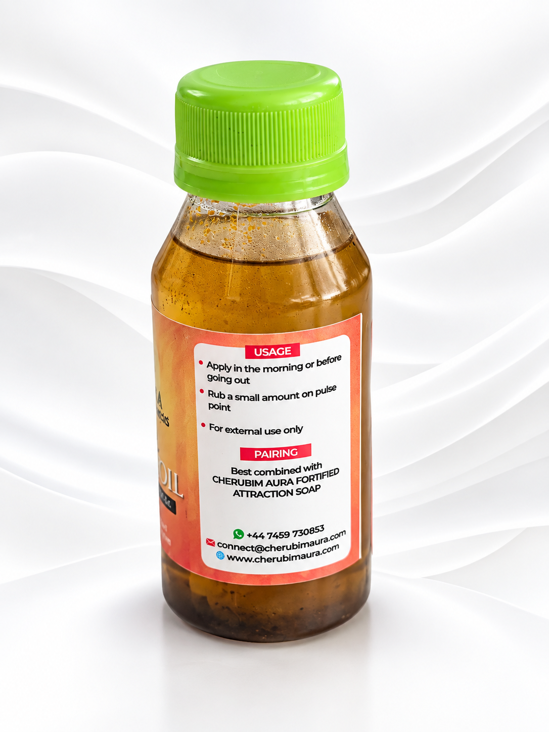 A small amber bottle with a bright green cap and a white label, photographed on a white background with soft wavy lines. The label includes usage instructions and contact information for Cherusm Aura Powered Attraction Soap.