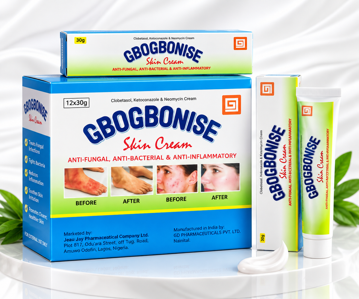 Gbogbonise Skin Cream product packaging and tubes arranged on a white reflective surface with a white draped backdrop. The set includes a large blue-and-green box, a smaller matching box, a tube, a single-use cream stick, and a dollop of white cream in the foreground.
