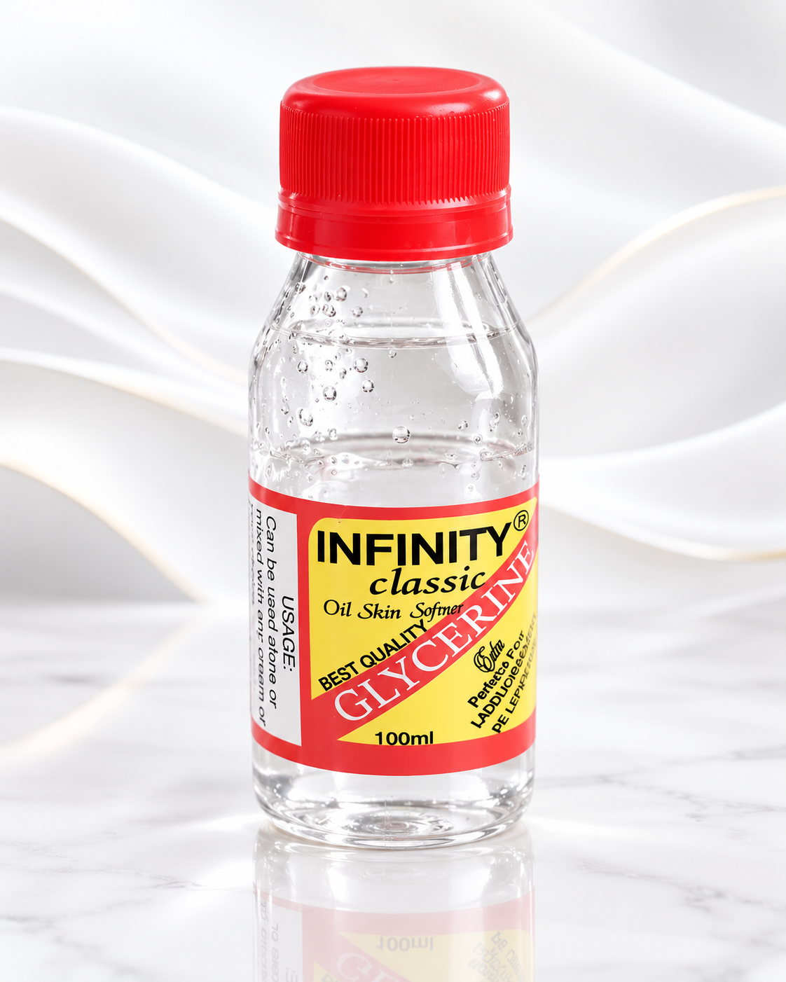 A 100ml clear plastic bottle of Infinity Classic Glycerine Oil Skin Softener with a red cap and a red, yellow, and black label, set against a white marble background with a soft white fabric drape.