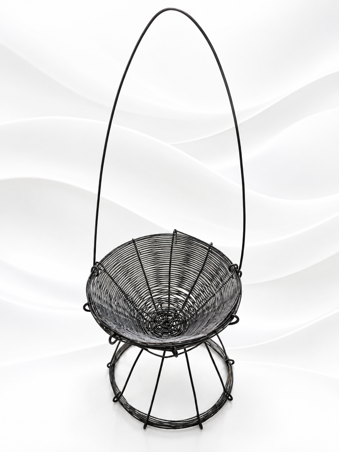 A black metal hanging basket chair with a woven seat and a tall arched handle, shown against a white wavy studio background.