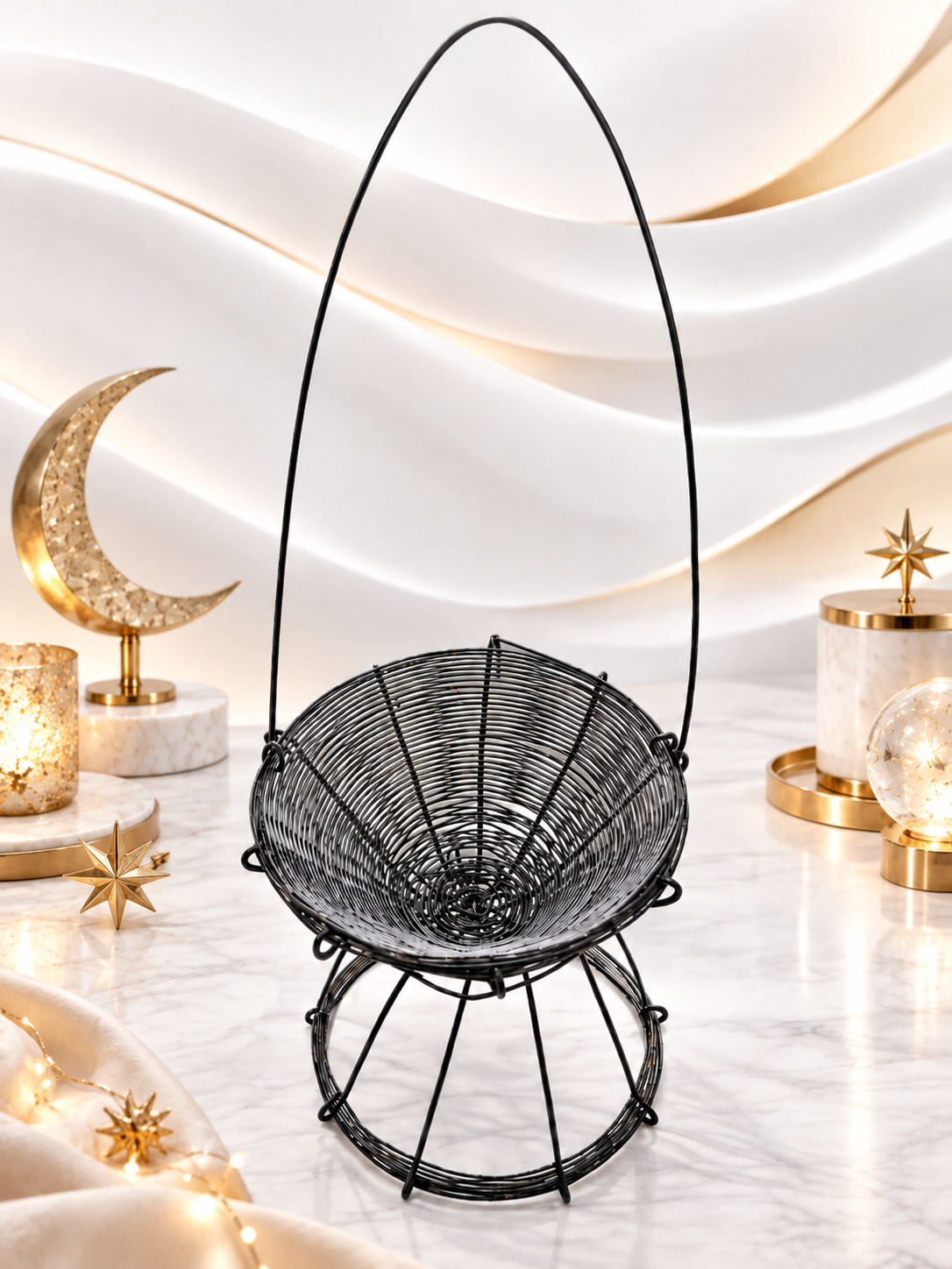 A black metal hanging basket with a woven circular body and tall arched handle, displayed on a marble surface with warm decorative lights and gold-toned ornaments in the background.