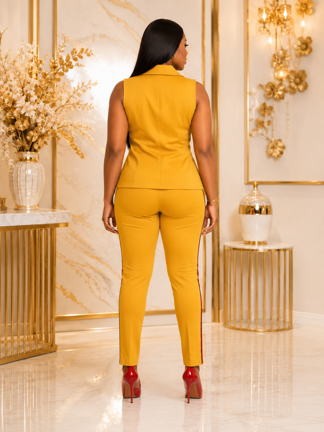 A woman shown from the back wearing a mustard yellow sleeveless blazer and matching slim-fit pants with red high heels, standing in a luxurious room with gold accents and marble walls.
