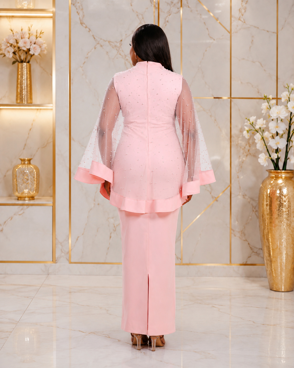 A woman viewed from behind wearing a light pink traditional outfit with sheer flutter sleeves and a matching long skirt, standing in a marble-finish room with gold-framed walls and decorative vases.