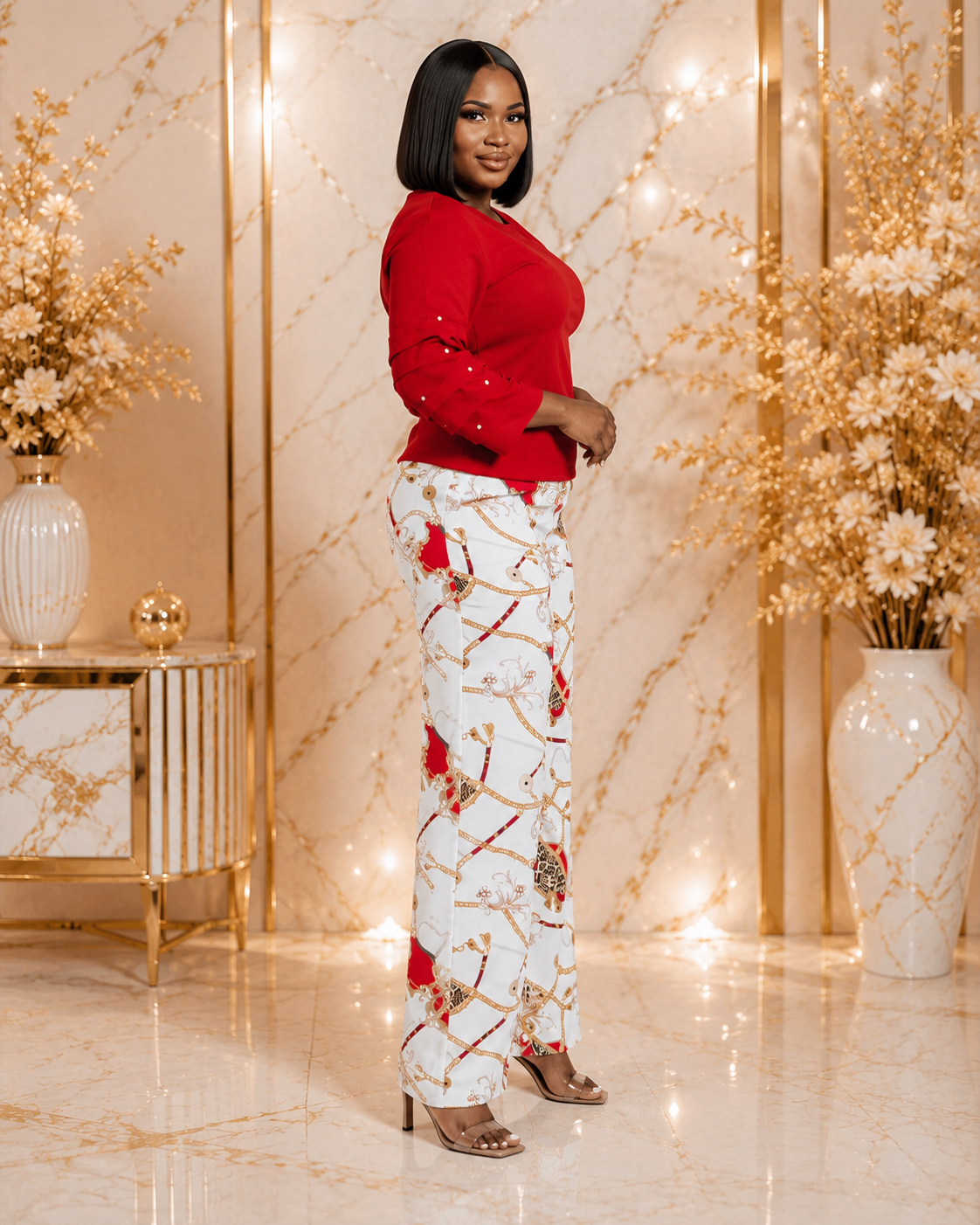 A woman stands in a decorated indoor setting wearing a bright red long-sleeve top and white wide-leg pants with a red, gold, and green floral pattern. The background features gold marble panels, warm string lights, and white floral arrangements.