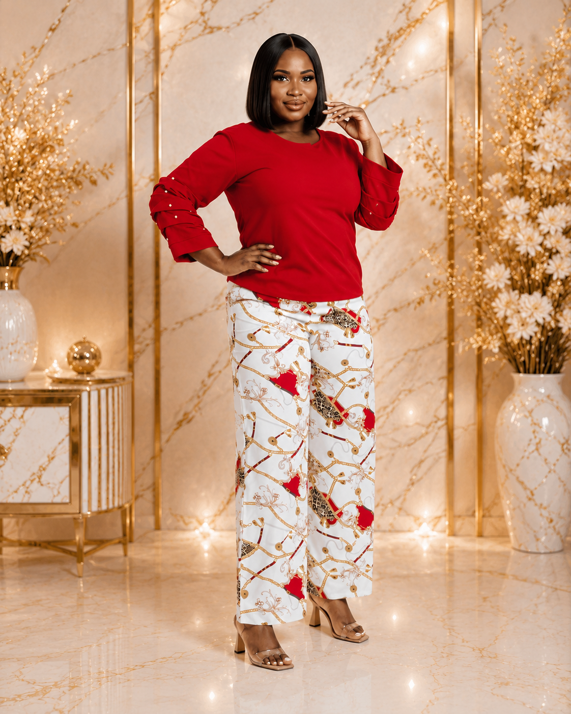 A woman modeling a red long-sleeve top and white wide-leg trousers with a red, gold, and brown abstract pattern, standing in a luxurious room with gold marble walls and decorative vases.
