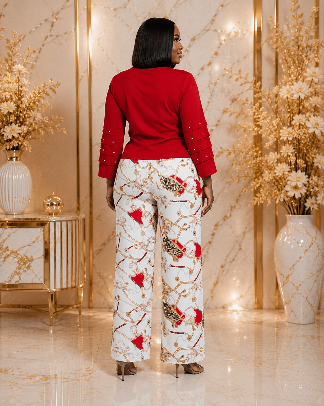 A woman shown from the back wearing a bright red long-sleeve top and white wide-leg pants with a red, gold, and brown abstract pattern, standing in a luxurious room with gold accents and marble floors.