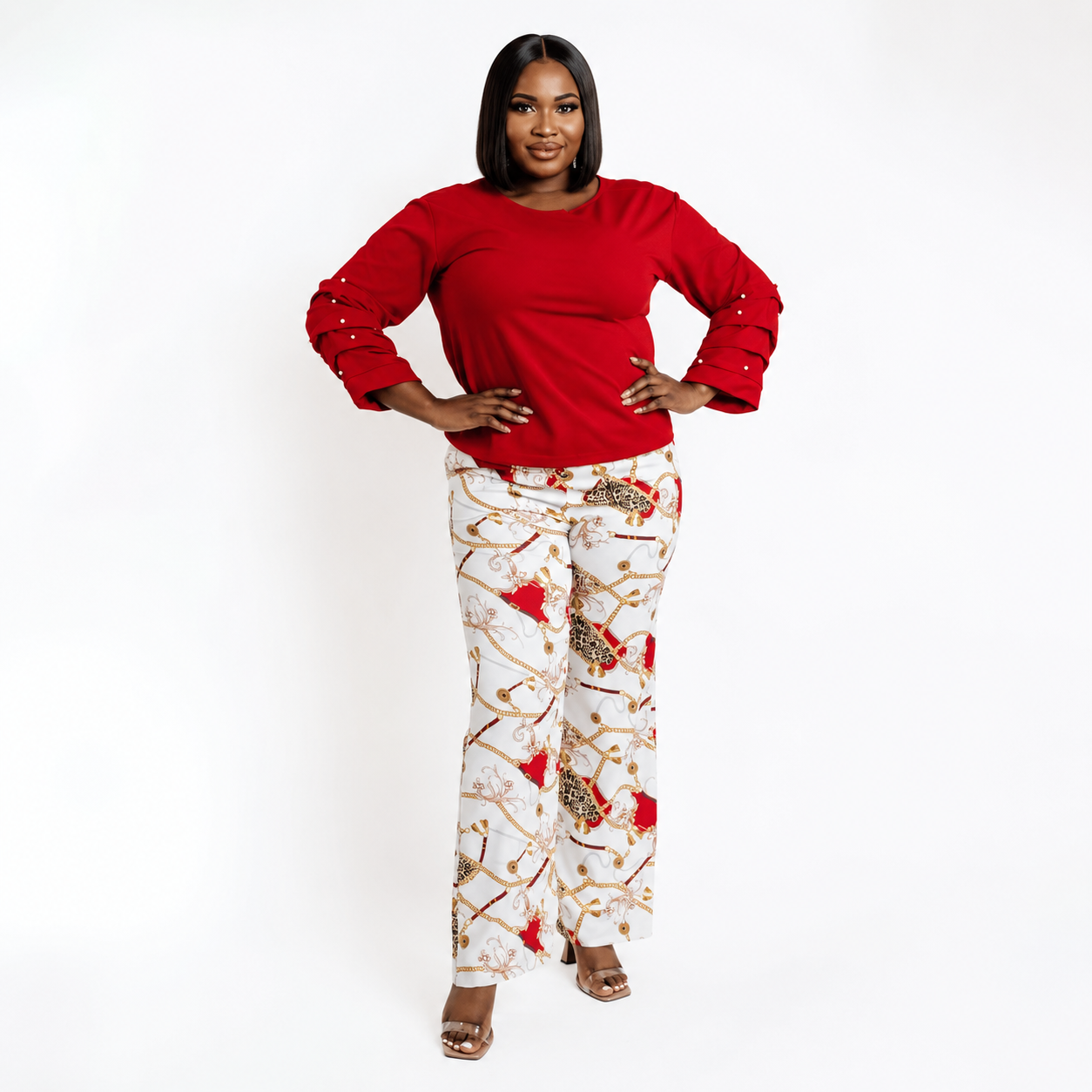 A woman modeling a red long-sleeve top and white pants with a red, gold, and brown abstract pattern, standing against a plain white studio background.
