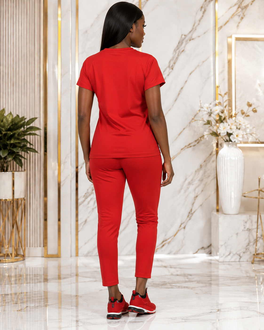 A woman stands with her back to the camera wearing a matching red short-sleeve top and slim-fit pants set, paired with red sneakers, in a luxury interior with marble walls and gold accents.