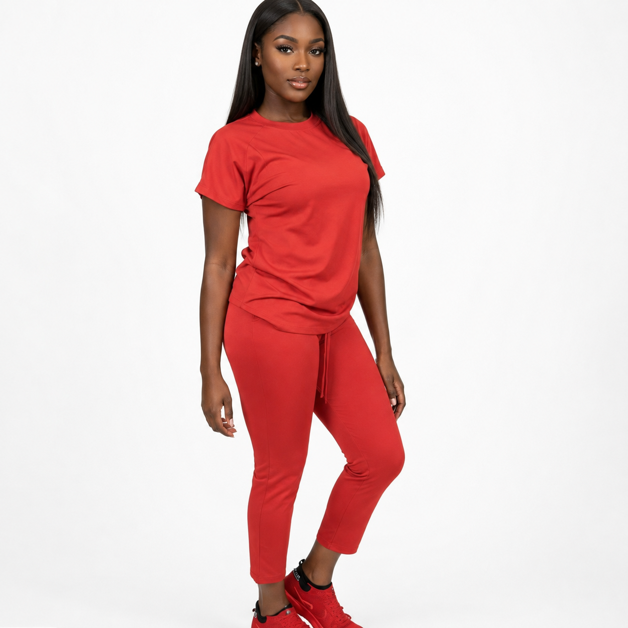 A woman modeling a matching red two-piece lounge set consisting of a short-sleeve crewneck t-shirt and cropped drawstring pants, paired with red sneakers, against a solid white background.