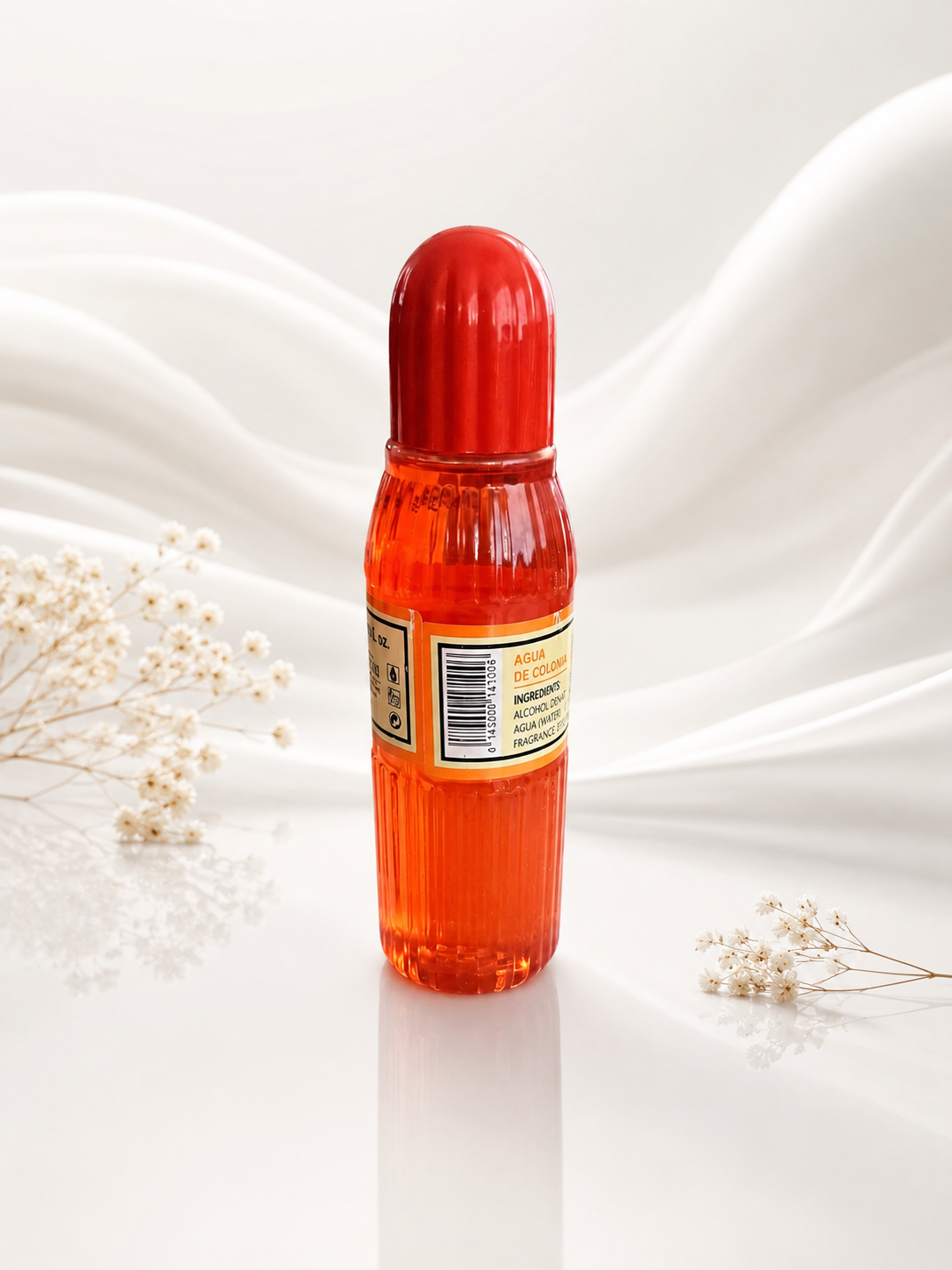 A ribbed clear bottle filled with bright orange-red liquid, topped with a glossy red cap, standing upright on a white surface with soft white fabric folds and small dried flowers around it.