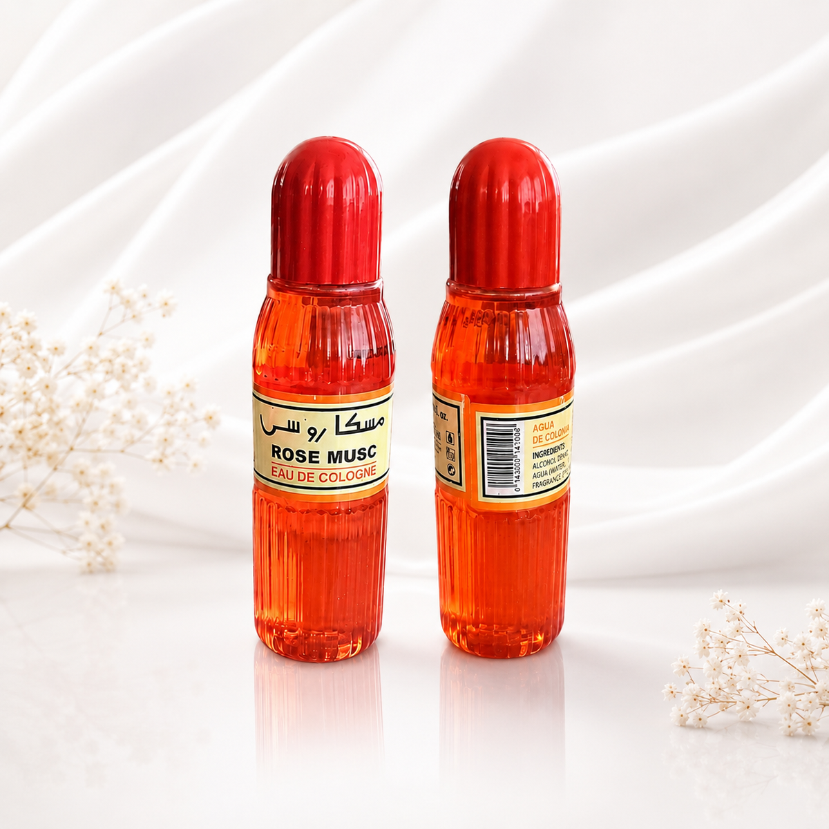 Two small ribbed glass perfume bottles filled with bright orange-red liquid, each with a red cap and a yellow-and-black label reading ROSE MUSC EAU DE COLLAR, set on a white surface against a white draped fabric background with small white flowers at the sides.
