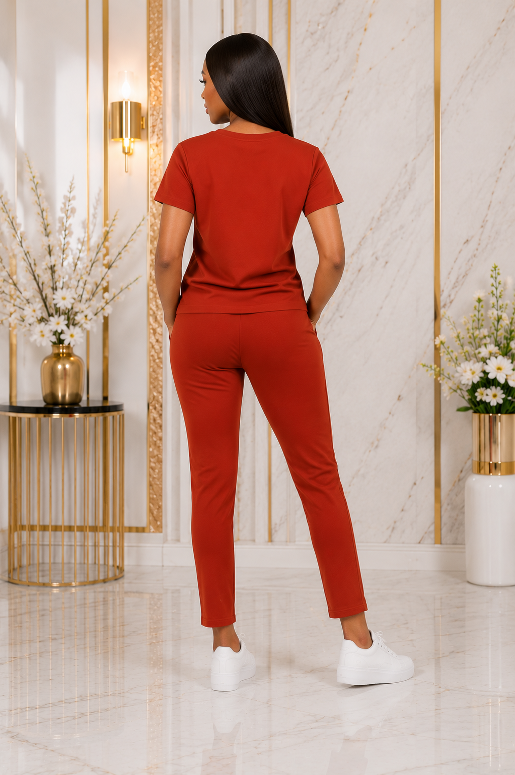 A woman stands with her back to the camera wearing a matching rust-orange short-sleeve top and slim-fit pants set, paired with white sneakers in a modern interior setting with marble walls and gold accents.