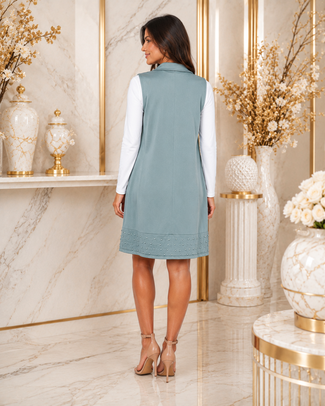 A woman wearing a sleeveless dusty blue dress over a white long-sleeve top, standing in a luxurious marble-floored room with gold accents and decorative vases.