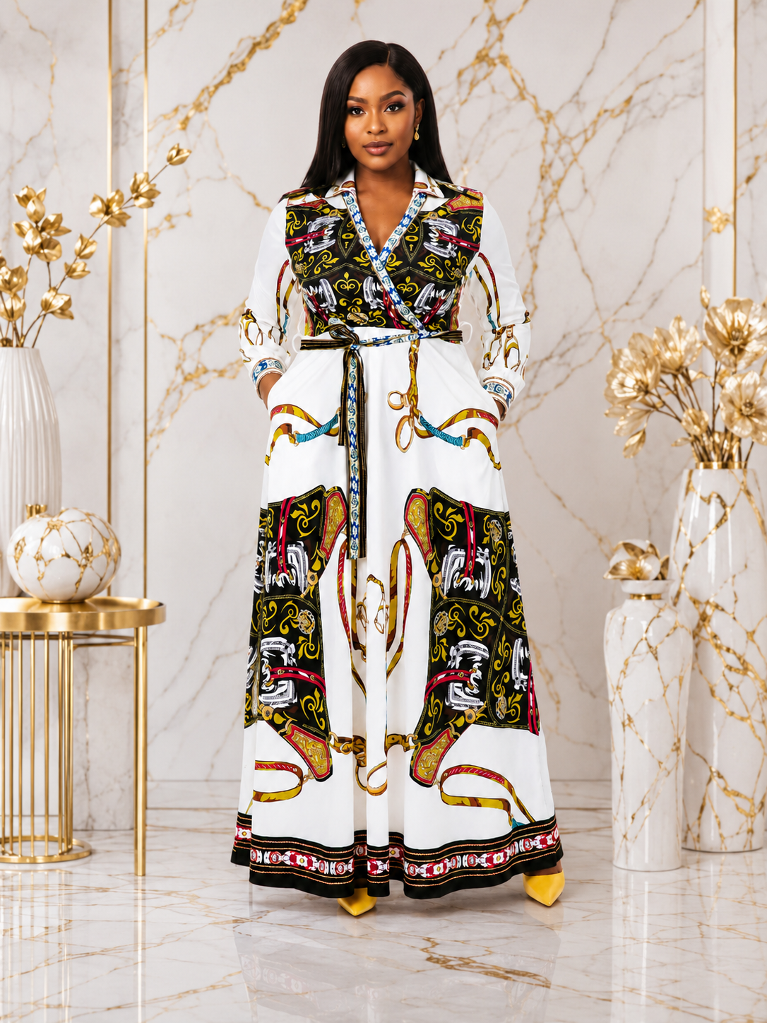 A woman modeling a long white maxi dress with a colorful, ornate graphic print featuring black, green, red, and gold elements, set against a marble wall with gold decorative accents.