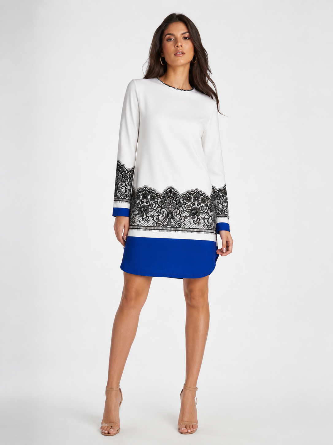 A woman modeling a long-sleeve white knit dress with a black damask pattern across the lower sleeves and hem, and a solid royal blue skirt section, set against a plain light gray studio background.