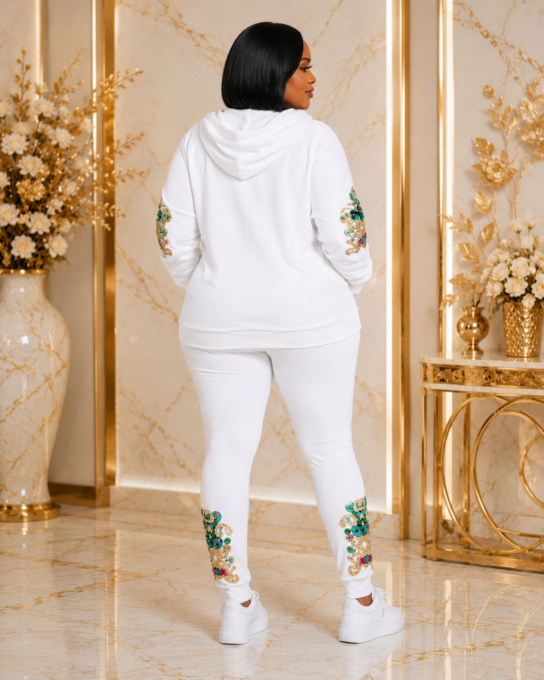 A woman stands with her back to the camera wearing a white two-piece loungewear set featuring floral embroidery on the sleeves and lower legs, set against a luxurious gold and beige marble interior.